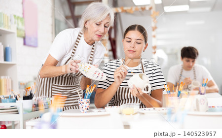 Senior woman assisting young girl painting ceramic mug in pottery class Senior woman assisting young girl painting ceramic mug in pottery class 140523957