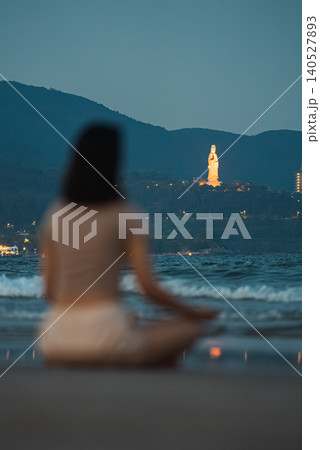 Woman meditating on beach shoreline with sea waves and Lady Buddha in Da Nang, Female practicing mindfulness by ocean at sunset with coastal mountains 140527893