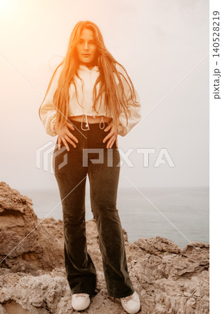 Woman standing cliffs posing on rocky terrain by the ocean with copy space and warm sunset lighting Woman standing cliffs posing on rocky terrain by the ocean with copy space and warm sunset lighting 140528219