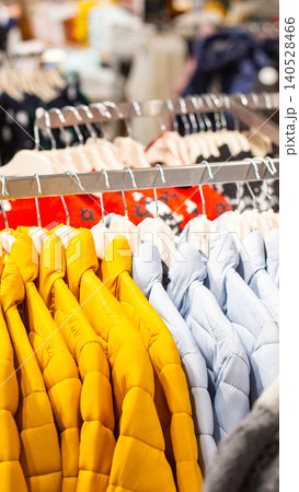 Rows of vibrant yellow and soft light blue padded puffer jackets hanging on display in a modern clothing store, showcasing new arrivals for cold weather, vertical photo 140528466