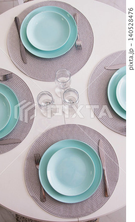 Elegant dining table set for three with aquamarine plates, grey woven placemats, and polished silverware, clear empty glasses on white surface, overhead view vertical photo Elegant dining table set for three with aquamarine plates, grey woven placemats, and polished silverware, clear empty glasses on white surface, overhead view vertical photo 140528476