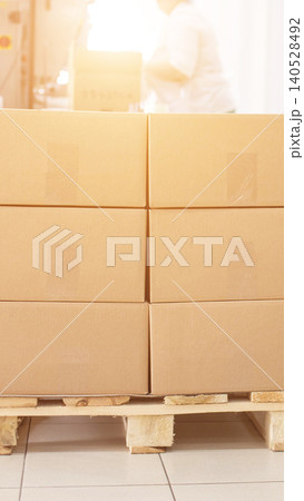 Neatly stacked brown cardboard boxes on sturdy wooden pallet ready for distribution or storage, production facility background with blurry worker, bright light flare vertical photo Neatly stacked brown cardboard boxes on sturdy wooden pallet ready for distribution or storage, production facility background with blurry worker, bright light flare vertical photo 140528492