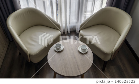 Top view of two cream leather armchairs with two white coffee cups on a round wooden table. Sheer curtains and dark floor in a cozy hotel room in Vietnam. Top view of two cream leather armchairs with two white coffee cups on a round wooden table. Sheer curtains and dark floor in a cozy hotel room in Vietnam. 140528522