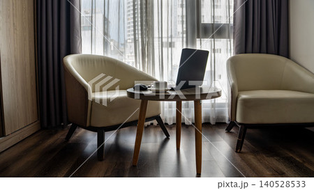 Cozy hotel room workspace with a laptop, coffee cup, and mouse on a round wooden table. Elegant cream armchair, sheer curtains, and city view in the background. Cozy hotel room workspace with a laptop, coffee cup, and mouse on a round wooden table. Elegant cream armchair, sheer curtains, and city view in the background. 140528533