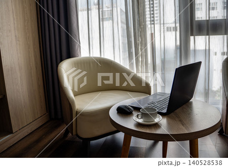 Cozy hotel room workspace with a laptop, coffee cup, and mouse on a round wooden table. Elegant cream armchair, sheer curtains, and city view in the background. 140528538