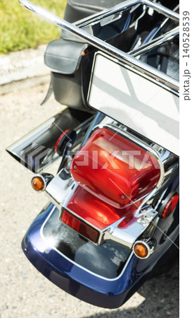Rear view of blue motorcycle parked on asphalt showing red taillights, amber turn signals, shining chrome luggage rack, and black leather saddlebag in vertical shot. 140528539