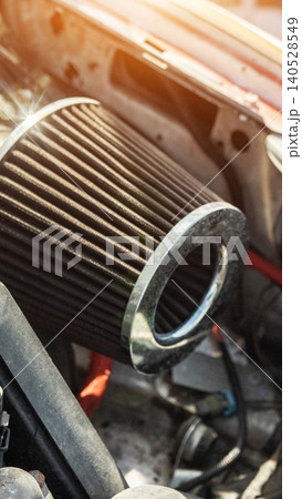 Performance air filter with metallic ring inside engine bay of modified vehicle, visible wires and hoses, bright lens flare from daylight, close up of tuning component, vertical orientation 140528549