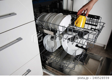 Human hand loading dirty dishes into modern automatic dishwasher kitchen appliance. White ceramic plates yellow mug organized inside kitchen dishwasher. 140528559