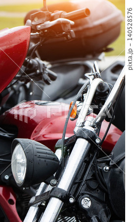 Close-up view of front part of red motorcycle with chrome handlebars and headlight, showing water droplets on surface, other parked motorcycles visible in background, vertical composition. Close-up view of front part of red motorcycle with chrome handlebars and headlight, showing water droplets on surface, other parked motorcycles visible in background, vertical composition. 140528576