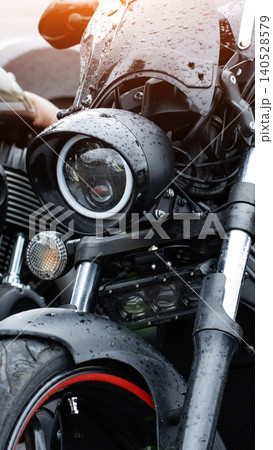 Close up front view of modern motorcycle with round LED headlight and rain droplets on windscreen and bodywork, detail of wet front wheel fender with red accent on rim, vehicle outdoors rainy day Close up front view of modern motorcycle with round LED headlight and rain droplets on windscreen and bodywork, detail of wet front wheel fender with red accent on rim, vehicle outdoors rainy day 140528579