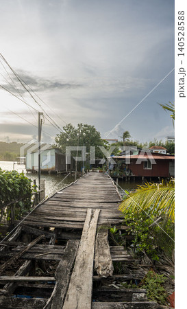 A crumbling wooden boardwalk leads through a remote Vietnamese fishing village with stilt houses, palm trees and power lines under a pale cloudy sky. Vietnam. A crumbling wooden boardwalk leads through a remote Vietnamese fishing village with stilt houses, palm trees and power lines under a pale cloudy sky. Vietnam. 140528588