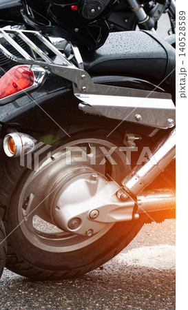 Black motorcycle rear wheel detail, wet from rain, with chrome exhaust pipes, fender, taillight, and turn signal, parked on asphalt, vertical shot. Black motorcycle rear wheel detail, wet from rain, with chrome exhaust pipes, fender, taillight, and turn signal, parked on asphalt, vertical shot. 140528589