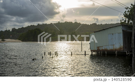 Golden sunlight breaks through storm clouds over a Vietnamese bay, shimmering on water beside a rusty corrugated stilt shed with old wooden poles. Vietnam. 140528590