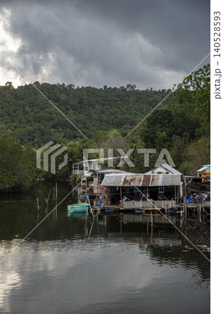 Rustic stilt houses on a calm Vietnamese river under dramatic storm clouds, with lush jungle hills and a small green boat reflected in still water. Vietnam. 140528593