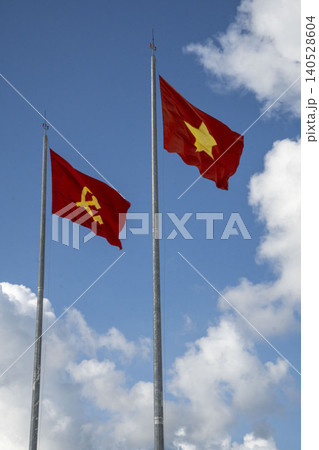 Vietnamese national flag and Communist Party flag wave side by side on tall flagpoles against a blue sky with white clouds in Vietnam. National patriotic symbols. Vietnamese national flag and Communist Party flag wave side by side on tall flagpoles against a blue sky with white clouds in Vietnam. National patriotic symbols. 140528604
