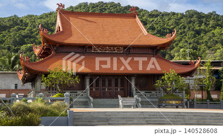 Side-angle view of the Ho Chi Minh memorial temple in Vietnam showing intricate wooden facade and sweeping traditional orange tiled roof against forested hills. Side-angle view of the Ho Chi Minh memorial temple in Vietnam showing intricate wooden facade and sweeping traditional orange tiled roof against forested hills. 140528608