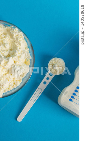 Close-up of dry infant formula powder in glass bowl with white measuring scoop, baby bottle alongside on vibrant blue background, preparation for infant feeding, vertical photo Close-up of dry infant formula powder in glass bowl with white measuring scoop, baby bottle alongside on vibrant blue background, preparation for infant feeding, vertical photo 140528618