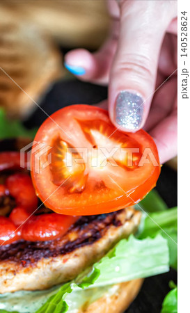 Woman hand placing fresh ripe tomato slice on grilled chicken sandwich with vibrant green lettuce and creamy sauce, preparing delicious meal close up vertical photo 140528624