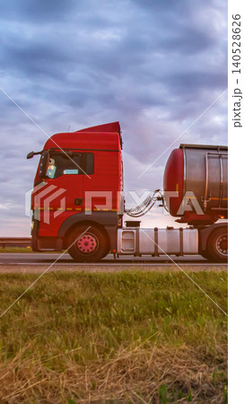 Red semi-truck with silver tanker trailer driving on highway against dramatic cloudy sky sunset, transport of liquid cargo on road, freight delivery logistics vertical photo 140528626