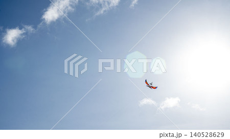 A small colorful kite flies high in a bright blue sky near the blazing sun with lens flare and white clouds. Carefree summer leisure moment in Vietnam. A small colorful kite flies high in a bright blue sky near the blazing sun with lens flare and white clouds. Carefree summer leisure moment in Vietnam. 140528629