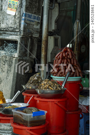 A Vietnamese street food stall with red plastic buckets used as improvised counters, displaying piles of dried sausages, pickled vegetables, and preserved foods in metal bowls. Vietnamese signage 140528661