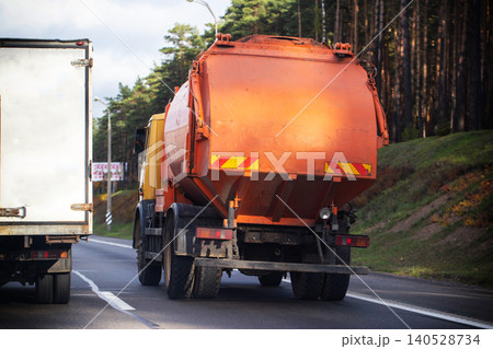 Rear view of orange garbage truck and white truck driving on asphalt road beside forest slope on overcast day Rear view of orange garbage truck and white truck driving on asphalt road beside forest slope on overcast day 140528734
