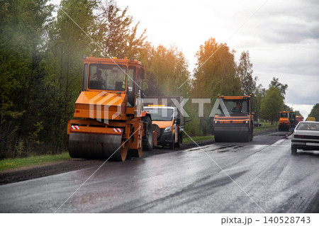 Orange road rollers compacting new asphalt surface during daytime road construction operations. Heavy machinery working on rural highway under cloudy sky. Copy space for text 140528743