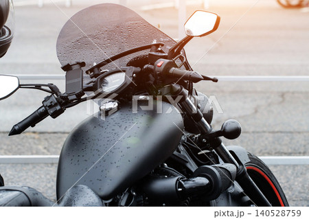 Black motorcycle front view close-up detail, raindrops on windshield and fuel tank, chrome accents, speedometer, handlebars, mirror, outdoor setting, after rain with sun flare 140528759