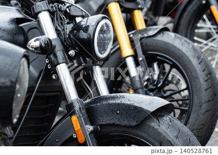 Close-up detail of front section of black motorcycle, featuring modern LED headlight, front fender, and chrome shock absorbers, surface covered with water drops after rain 140528761
