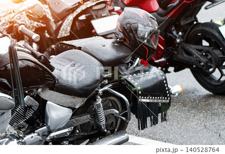 Black motorcycle with full-face helmet and studded leather saddlebags parked outdoors, covered in raindrops, after rain shower, detail of vehicle and gear, pavement surface, bright light effect Black motorcycle with full-face helmet and studded leather saddlebags parked outdoors, covered in raindrops, after rain shower, detail of vehicle and gear, pavement surface, bright light effect 140528764