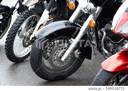 Close-up view of multiple motorcycles, including a cruiser, dirt bike, and sport bike, parked on wet asphalt after rain, showing wheels, fenders, engines, and chrome details with water droplets. 140528771