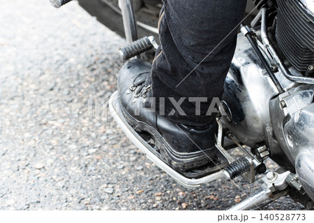 Rider foot black boot on motorcycle footboard, close-up shot showing leg and engine section, elements of road and biker machine visible, detail of chrome and dark metal parts, modern chopper style, Rider foot black boot on motorcycle footboard, close-up shot showing leg and engine section, elements of road and biker machine visible, detail of chrome and dark metal parts, modern chopper style, 140528773