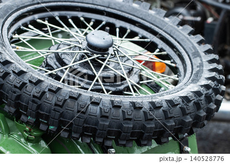 Close-up of wet knobby tire and spoke wheel of vintage motorcycle spare, showcasing water droplets on rugged rubber tread and shiny metal spokes, parked outdoors on rainy day Close-up of wet knobby tire and spoke wheel of vintage motorcycle spare, showcasing water droplets on rugged rubber tread and shiny metal spokes, parked outdoors on rainy day 140528776
