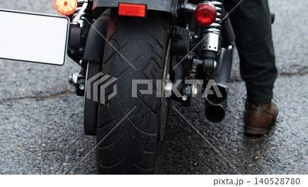 Rear view of motorcycle parked on wet asphalt road with person leg and brown shoe standing next to dual exhaust pipes and tail lights, license plate frame visible 140528780