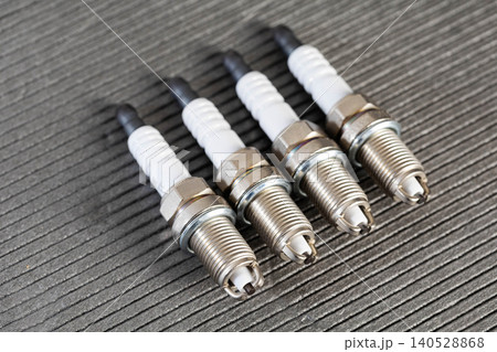 Four new automotive spark plugs for internal combustion engine, metallic thread and white ceramic insulator, arranged diagonally on dark textured surface. Four new automotive spark plugs for internal combustion engine, metallic thread and white ceramic insulator, arranged diagonally on dark textured surface. 140528868