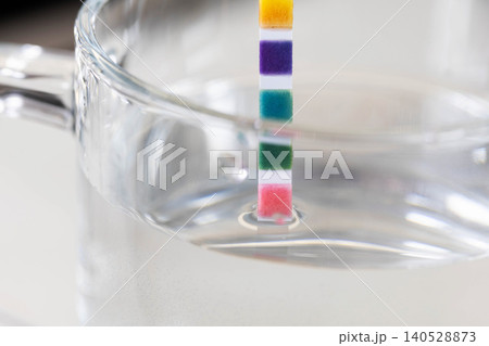 Colorimetric test strip immersed in clear water within glass beaker, indicating various water quality parameters through color change, showing chemical reaction process for analysis Colorimetric test strip immersed in clear water within glass beaker, indicating various water quality parameters through color change, showing chemical reaction process for analysis 140528873