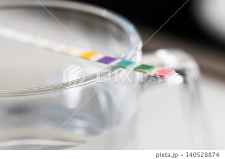 Colored test strip partially submerged in clear liquid of glass container, indicating water parameters measurement with visible chemical reaction squares, close up view. Colored test strip partially submerged in clear liquid of glass container, indicating water parameters measurement with visible chemical reaction squares, close up view. 140528874