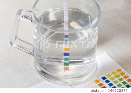 Process of water quality measurement shows a multi-parameter test strip dipped in clear glass of drinking water, with color comparison chart for accurate interpretation visible on a light-colored 140528875
