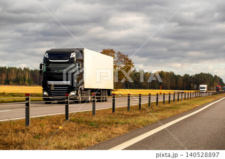 Black semi-truck with white trailer transports cargo on highway during autumn day under cloudy sky, movement blur conveys speed of transportation logistics Black semi-truck with white trailer transports cargo on highway during autumn day under cloudy sky, movement blur conveys speed of transportation logistics 140528897