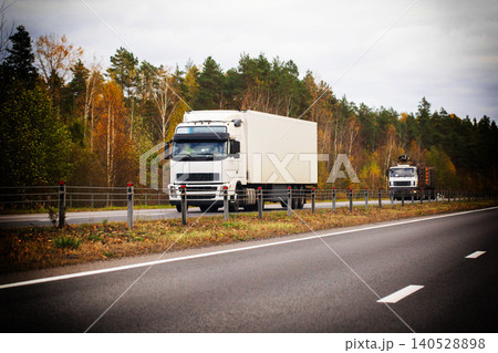 White semi-truck with cargo container drives on highway road during autumn season, logging truck follows, forest trees on roadside with fall foliage color White semi-truck with cargo container drives on highway road during autumn season, logging truck follows, forest trees on roadside with fall foliage color 140528898
