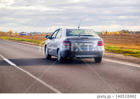 Silver sedan car driving away on rural asphalt road through countryside under cloudy sky, fields with dry grass and autumn trees along road Silver sedan car driving away on rural asphalt road through countryside under cloudy sky, fields with dry grass and autumn trees along road 140528902