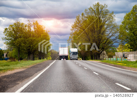 Two cargo trucks traveling on asphalt road through rural landscape under overcast sky, green trees lining route, transport vehicle movement on country highway. 140528927