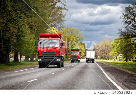 Red dump truck travels on rural road with green trees lining sides under cloudy sky, followed by other trucks in countryside landscape 140528928