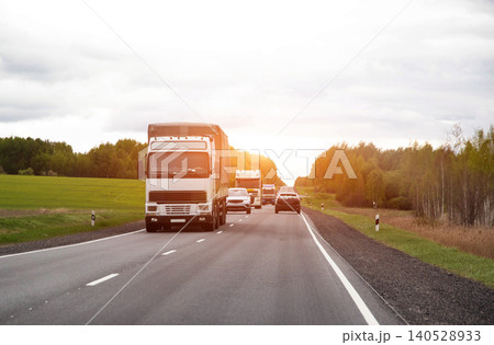 Traffic flow of commercial trucks and passenger cars on asphalt road through rural landscape under overcast sky with sunlight flare Traffic flow of commercial trucks and passenger cars on asphalt road through rural landscape under overcast sky with sunlight flare 140528933