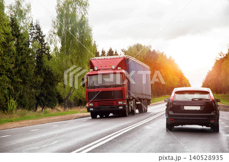 Red cargo truck with blue tarpaulin trailer and black passenger car traveling on wet country road surrounded by lush green trees on a cloudy day. 140528935