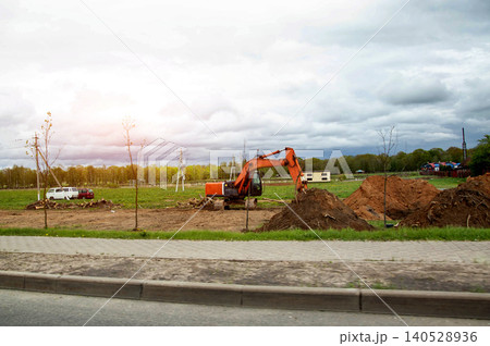Orange excavator performs earthwork at construction site with piles of dirt under cloudy sky. Green field, trees, and power poles visible in background 140528936