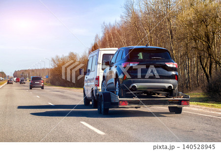 White cargo van transports black SUV on flatbed trailer, traveling on multi-lane highway with distant vehicles and bare trees under clear blue sky. White cargo van transports black SUV on flatbed trailer, traveling on multi-lane highway with distant vehicles and bare trees under clear blue sky. 140528945