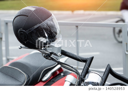 Black motorcycle helmet adorned with numerous raindrops, securely resting on handlebars of powerful red motorcycle, parked outdoors under soft ambient light, showcasing elements of modern biker Black motorcycle helmet adorned with numerous raindrops, securely resting on handlebars of powerful red motorcycle, parked outdoors under soft ambient light, showcasing elements of modern biker 140528953