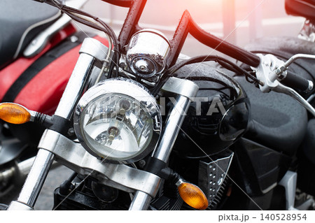 Close-up view of a wet black motorcycle front section with chrome headlight, handlebars, and signal lights, covered in raindrops after rain, showcasing cruiser style and robust engineering Close-up view of a wet black motorcycle front section with chrome headlight, handlebars, and signal lights, covered in raindrops after rain, showcasing cruiser style and robust engineering 140528954