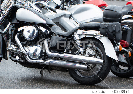 Close-up detail of modern cruiser motorcycle parked on wet asphalt surface on rainy day, chrome accents reflecting light, alongside other motorcycles and a black helmet, showcasing biker culture Close-up detail of modern cruiser motorcycle parked on wet asphalt surface on rainy day, chrome accents reflecting light, alongside other motorcycles and a black helmet, showcasing biker culture 140528956